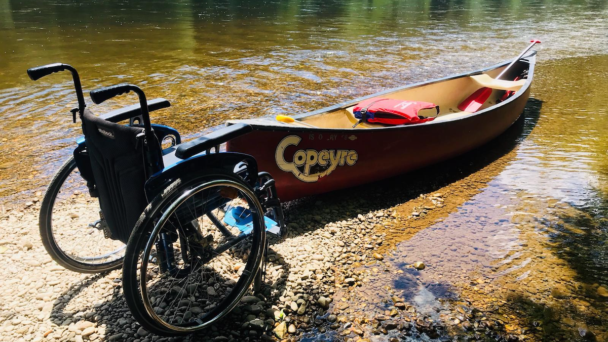 Ein Rollstuhl der an einem Kanu steht. Das Kanu liegt in der Dordogne. Regelmässig organisieren Arne und Silke vom le Colombie barrierefreie Kanutouren für die Gäste.